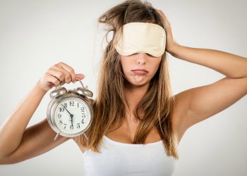 Beautiful young woman with sleeping mask holding alarm clock. She is tired and lazy in morning.