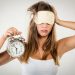 Beautiful young woman with sleeping mask holding alarm clock. She is tired and lazy in morning.